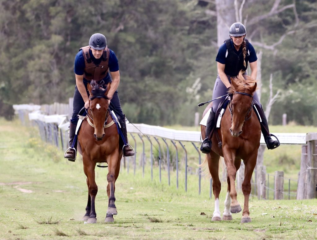 Two people riding brown horses on a grassy path, wearing helmets and casual riding gear. The person on the left is wearing a protective vest. Trees and a fence line the background. The scene appears relaxed and outdoorsy.