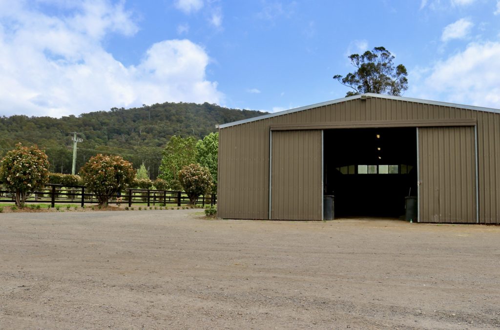 A large, open-sided barn stands on a dirt road, surrounded by greenery. In the background is a wooded hill under a partly cloudy blue sky. There are bushes and a wooden fence to the left of the barn.