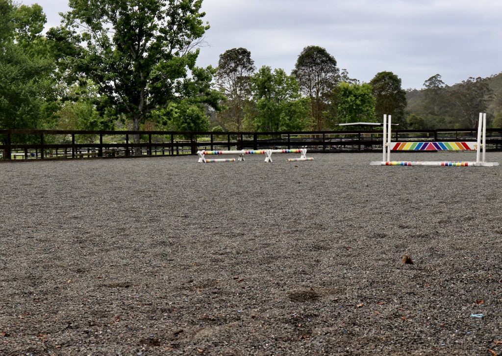 Outdoor equestrian arena with gravel surface, surrounded by trees and fencing. Colorful horse jumps are set up on the arena floor, including a striped bar jump. Overcast sky in the background.